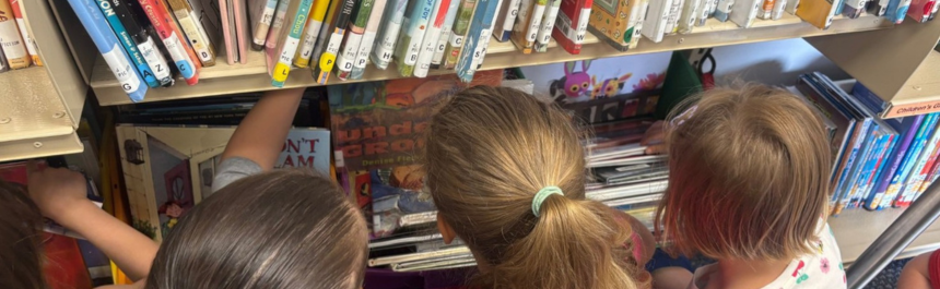 Text: three kids look at a bookshelf.