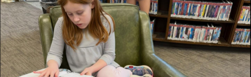 A child sits in a child-size soft chair and reads a book in a public library]