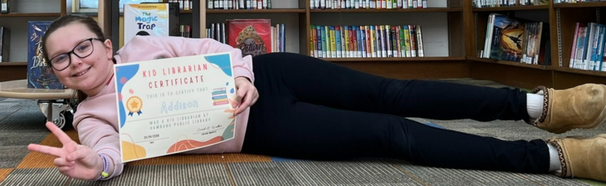 Child laying on the floor giving the peace sign as she holds her certificate naming her Kid Librarian for the Month